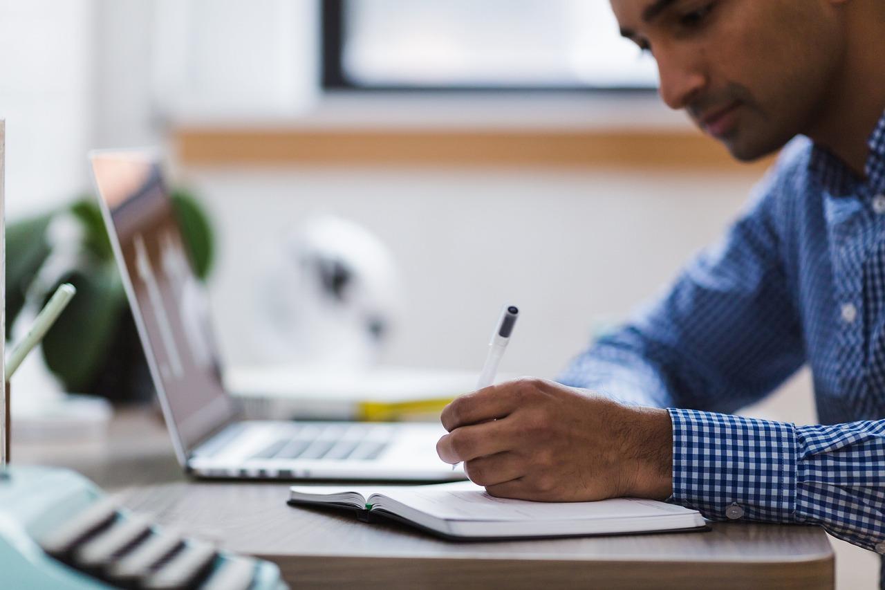 Homme écrivant des notes sur un bloc-notes à partir d'un ordinateur portable.