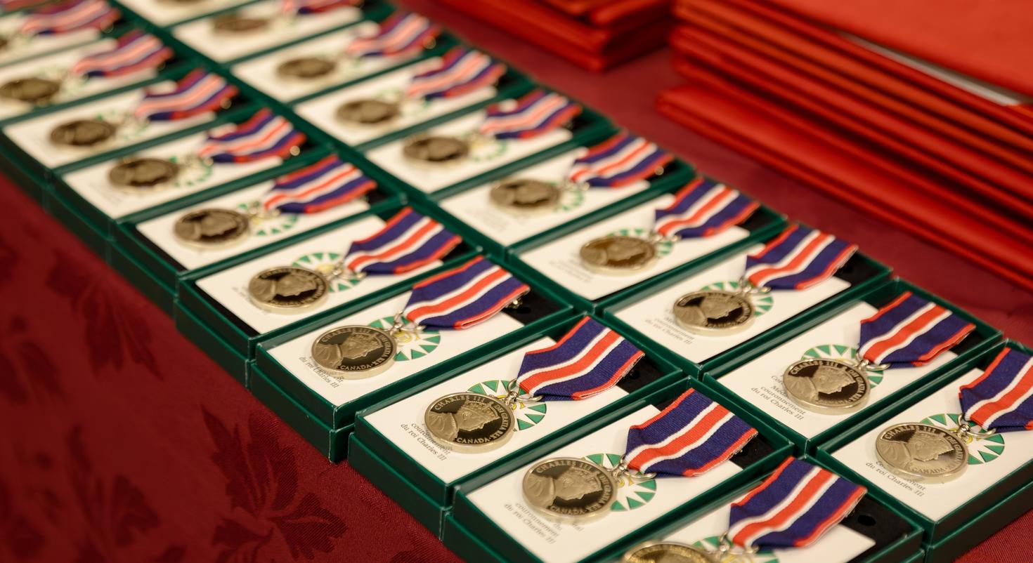 Plusieurs Médailles du couronnement du roi Charles III sur un présentoir. La médaille est circulaire, de couleur argent et attachée à un ruban bleu foncé, rouge vif et blanc.