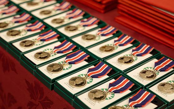 Plusieurs Médailles du couronnement du roi Charles III sur un présentoir. La médaille est circulaire, de couleur argent et attachée à un ruban bleu foncé, rouge vif et blanc.