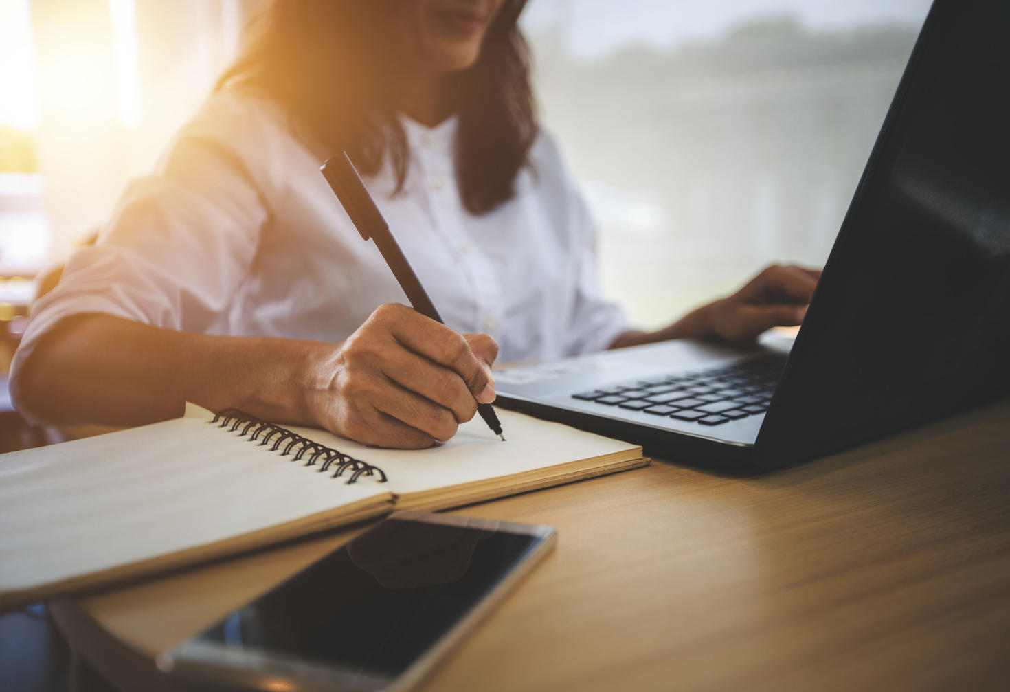 Personne travaillant à un bureau avec un ordinateur portable, prenant des notes dans un cahier à spirale, avec un téléphone intelligent placé à proximité.