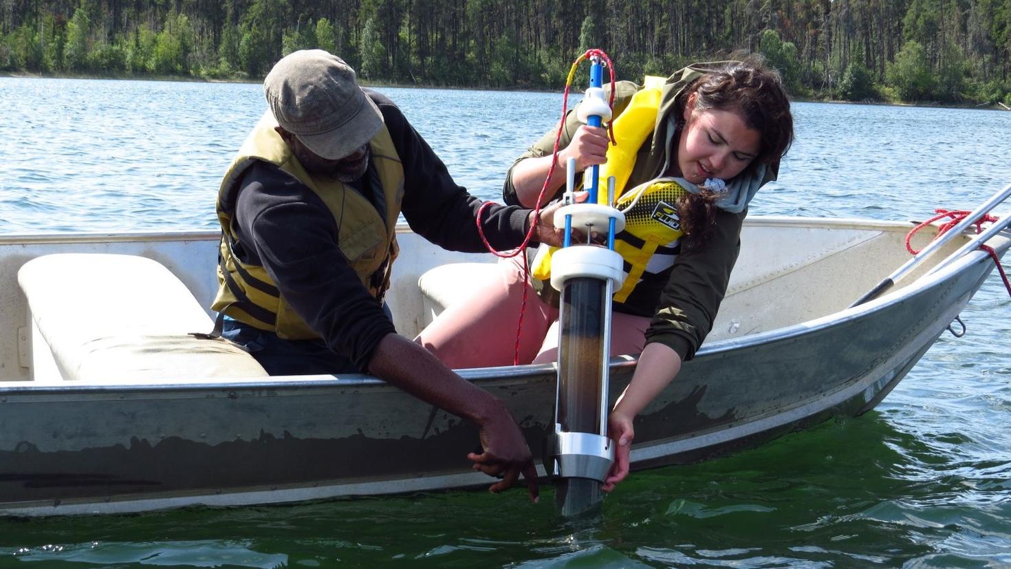 In a small boat, a research partner (left) and Dënë Cheecham-Uhrich (right) work together to pull up a sediment core.