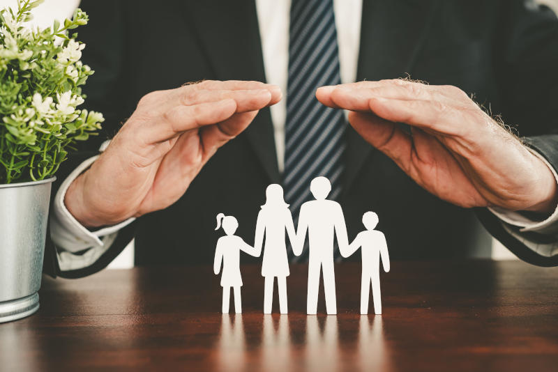 Une personne en tenue professionnelle tient ses mains en signe de protection au-dessus d'une silhouette en papier représentant une famille de quatre personnes se tenant par la main