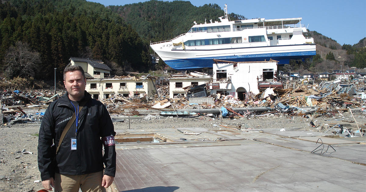 Le professeur Ioan Nestor se tient devant un bâtiment et un navire ravagés par le tsunami survenu en 2011 dans la région de Tōhoku au Japon.  