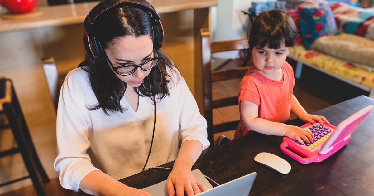 A woman working on her laptop as her young daughter sits beside her, playing on a toy computer.