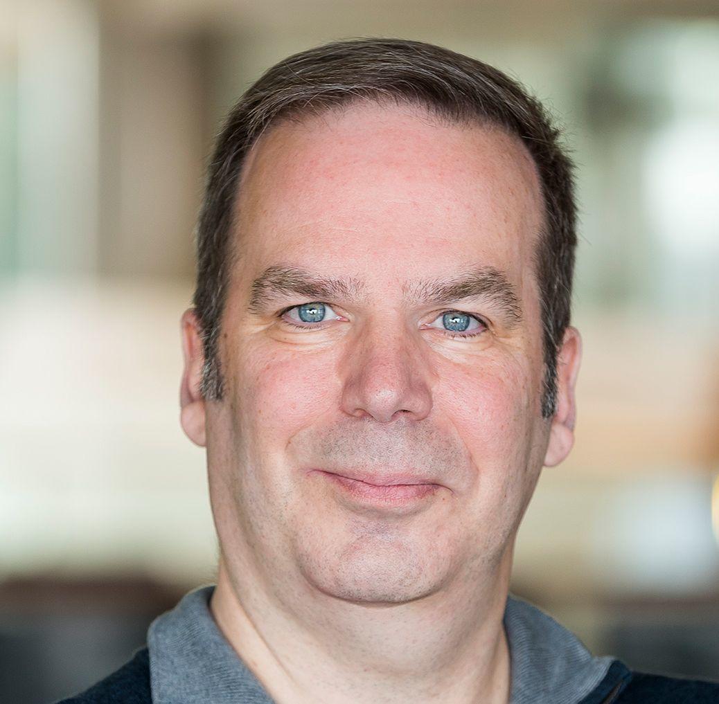 A man with short brown hair and bright blue eyes is shown in a close-up portrait