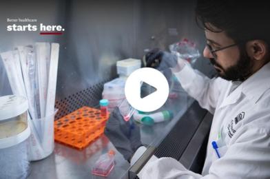 A healthcare researcher in a lab coat works inside a laboratory biosafety cabinet, handling samples with precision tools.