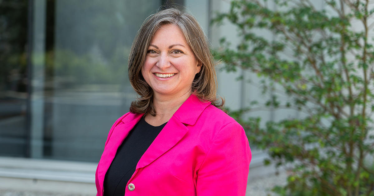 A person wearing a bright pink blazer over a black top stands outdoors . They are in front of a glass building with greenery in the background.