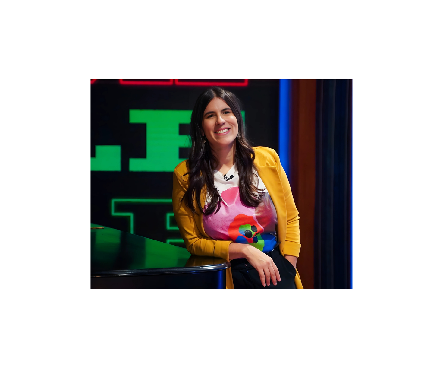 A person with long dark hair is smiling and leaning on a black surface. They are wearing a yellow jacket and a colorful t-shirt.