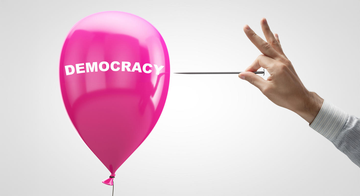 A pink balloon with the word "DEMOCRACY" written on it, being approached by a hand holding a needle, ready to pop it.