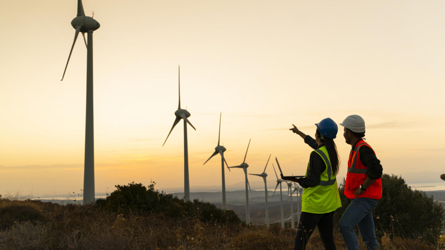Two engineers in safety gear inspect a wind farm at sunset, with one pointing towards a turbine while holding a laptop.