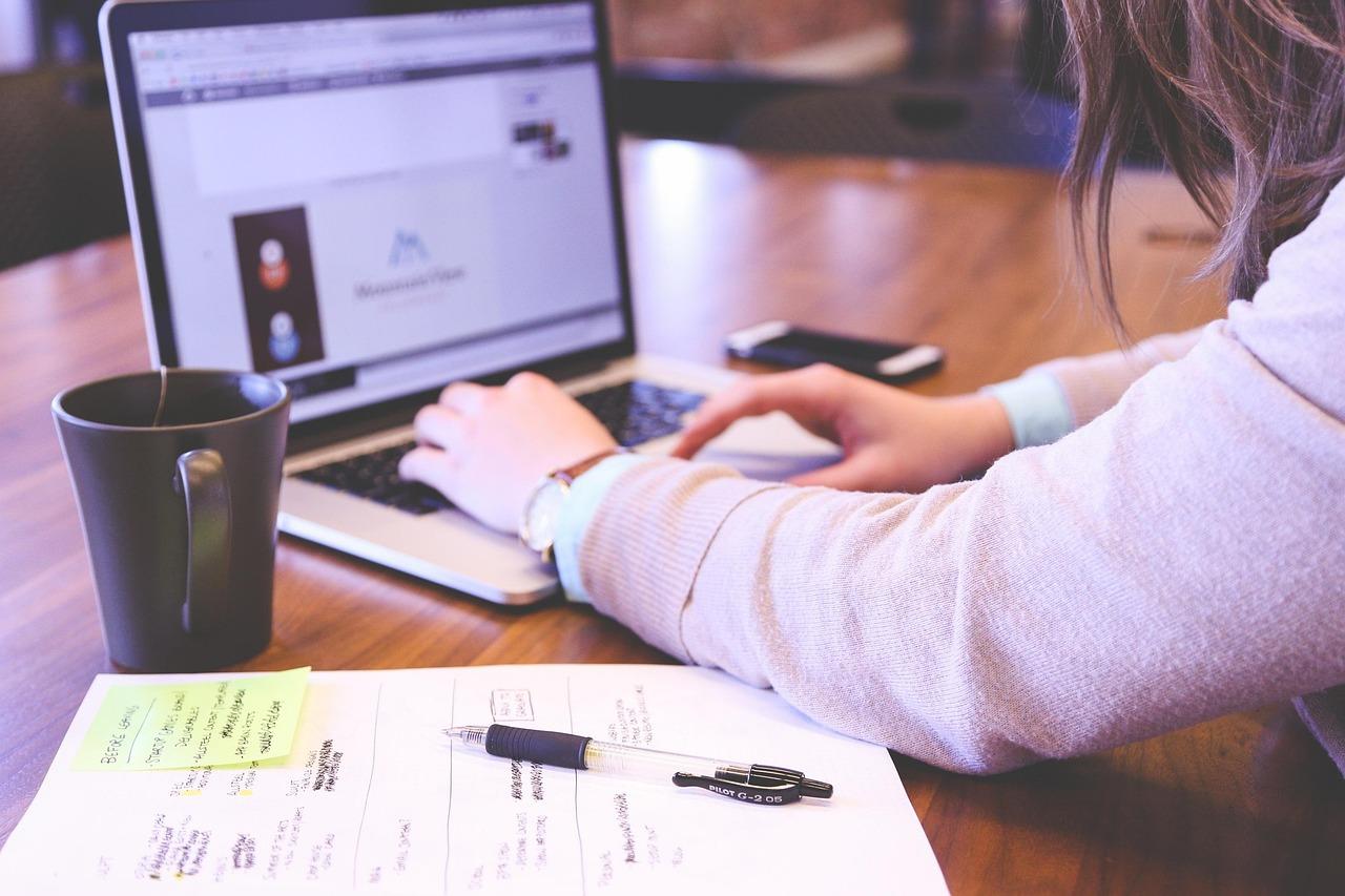 Woman typing on laptop keyboard with a notebook and coffee cup.