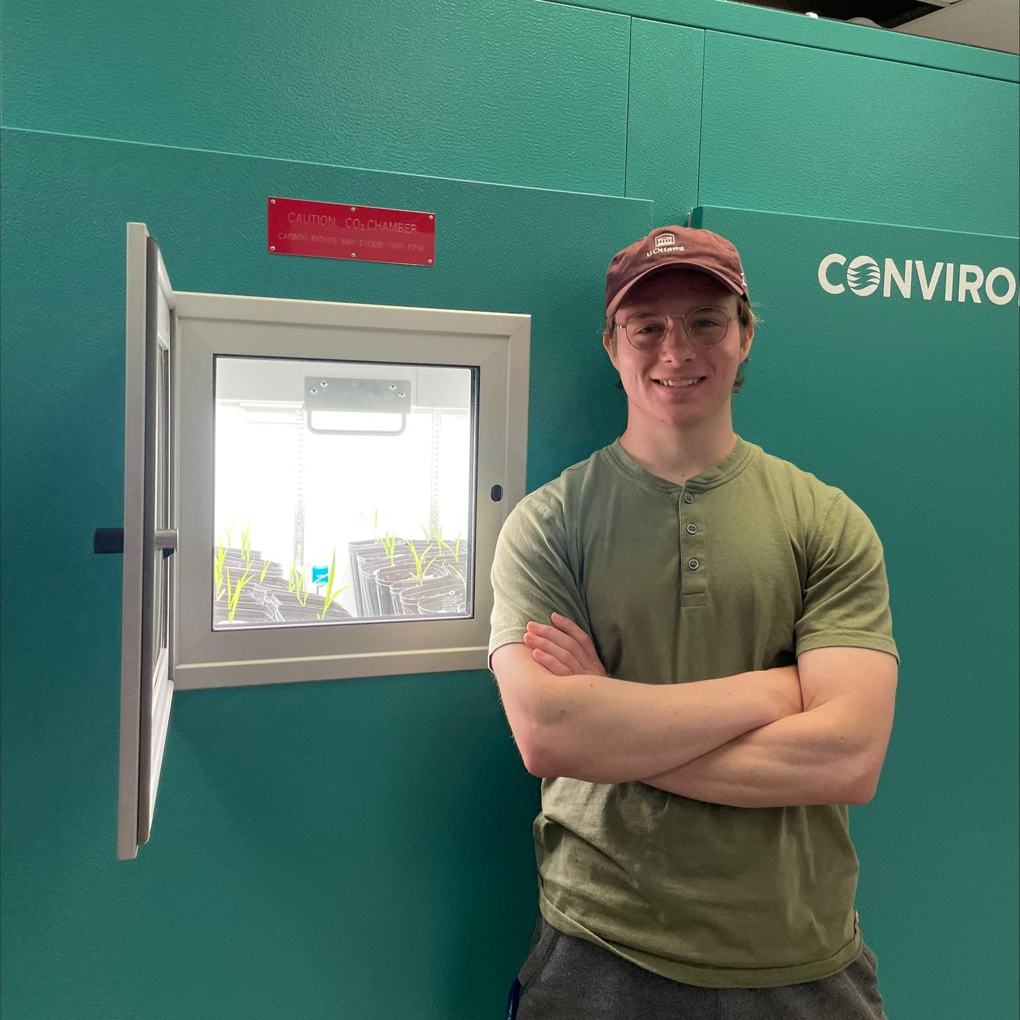 Robert Ferguson, who is a PhD student and MITACs intern, stands next to a growth chamber filled with Sudan grass inoculated with different strains of AMF.
