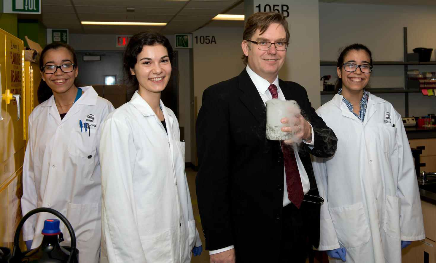 Professor Robert Pezacki stands in a laboratory holding a beaker filled with dry ice that is emitting visible vapor. He is flanked by three smiling PhD graduates in lab coats: Noreen Ahmed (far left), Roxana Filip (left of Pezacki), and Nadine Ahmed (far right). Laboratory equipment and signage are visible in the background.