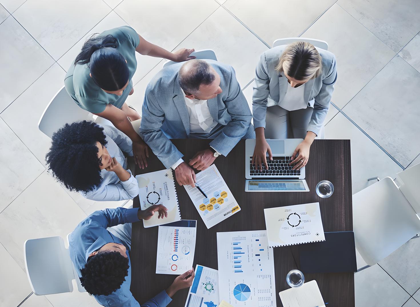 Group of professionals gathered around a table reviewing charts, graphs, and a laptop during a business meeting.