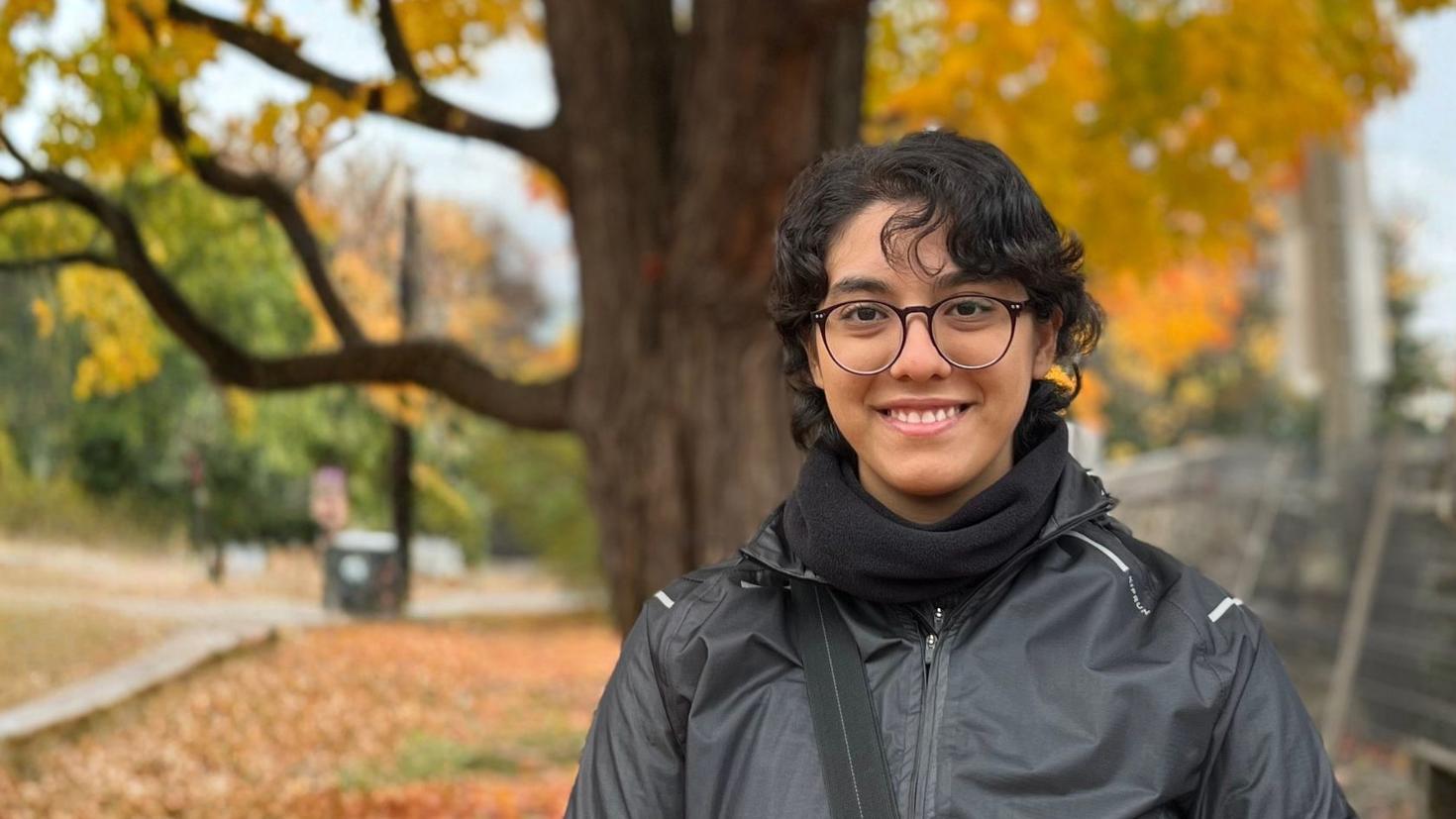 Nazanin Dehghan stands outdoors in front of a large tree with vibrant yellow and orange autumn leaves. She is smiling and wearing glasses, a black scarf and a dark windbreaker. The background is a slightly blurred urban setting with fallen leaves on the ground.