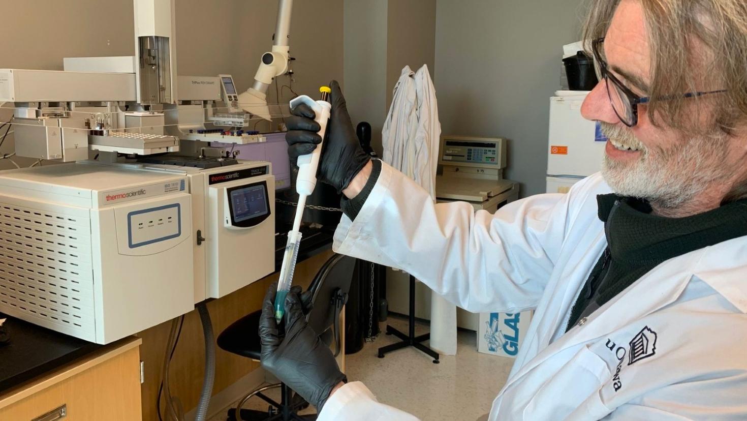 Professor Cory Harris, wearing a white lab coat and black gloves uses a pipette in the laboratory.