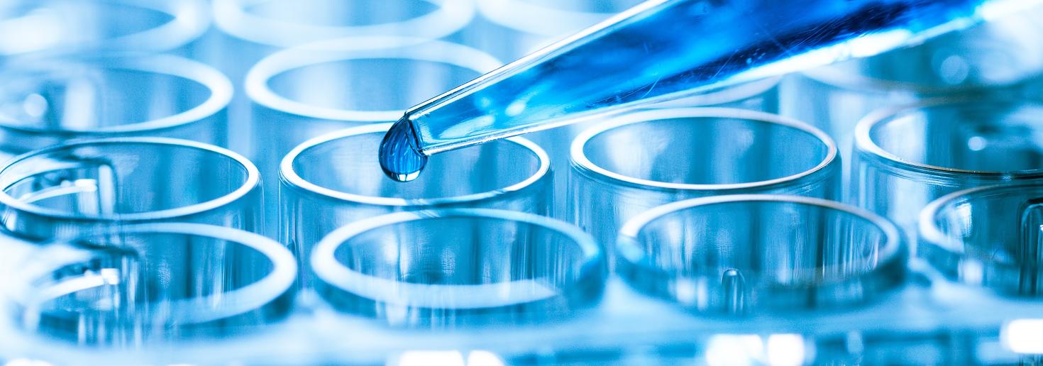 Close-up of a laboratory pipette dispensing a drop of liquid into a glass test tube among a rack of test tubes.