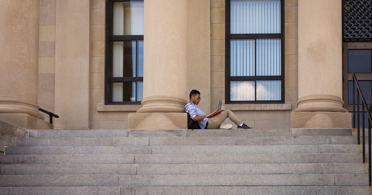 Un étudiant avec son ordinateur portable assis seul sur les marches du pavillon Tabaret.