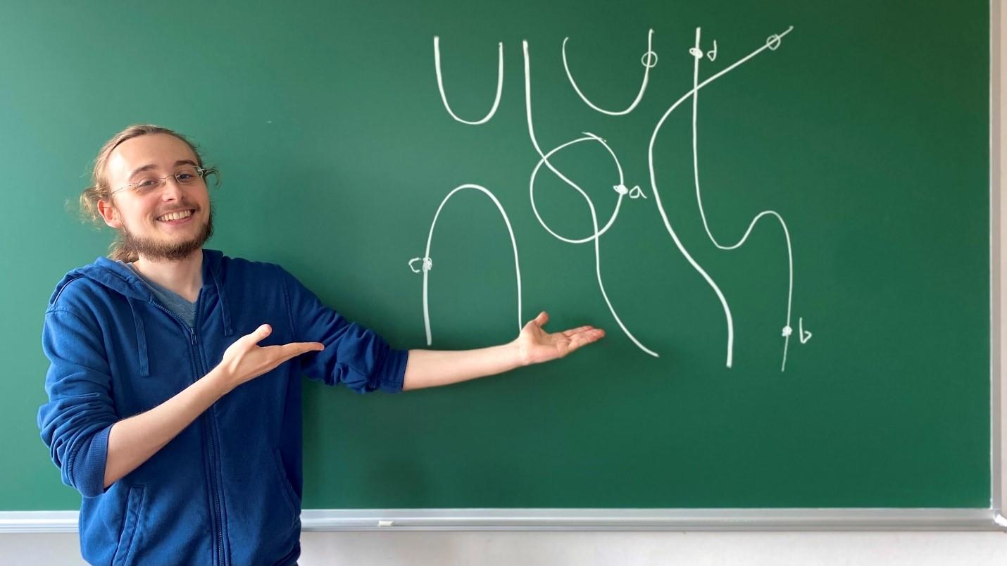 Master’s graduate Saima Samchuck-Schnarch wearing a blue hoodie and displaying Brauer diagrams illustrated on a green chalkboard.