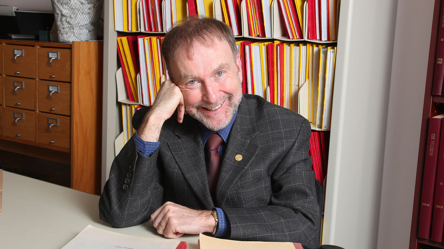 Michel Prévost assis à son bureau devant une bibliothèque chargée de livres.