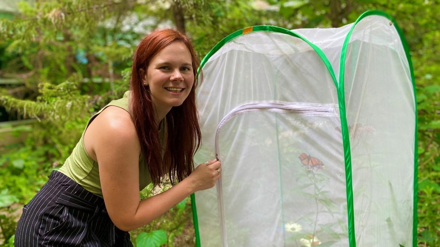 L’étudiante au doctorat Megan Reich porte un haut vert sans manches et un pantalon rayé noir et blanc. Elle pose en plein air, à côté d'une tente en filet blanc contenant des plantes et des papillons monarques.