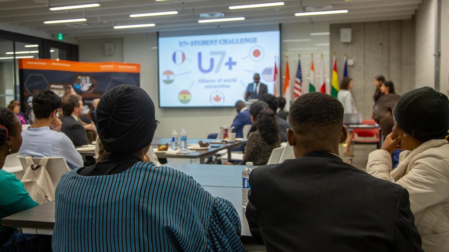 Les personnes assistant à la cérémonie et regardant l'écran affichant le logo U7+