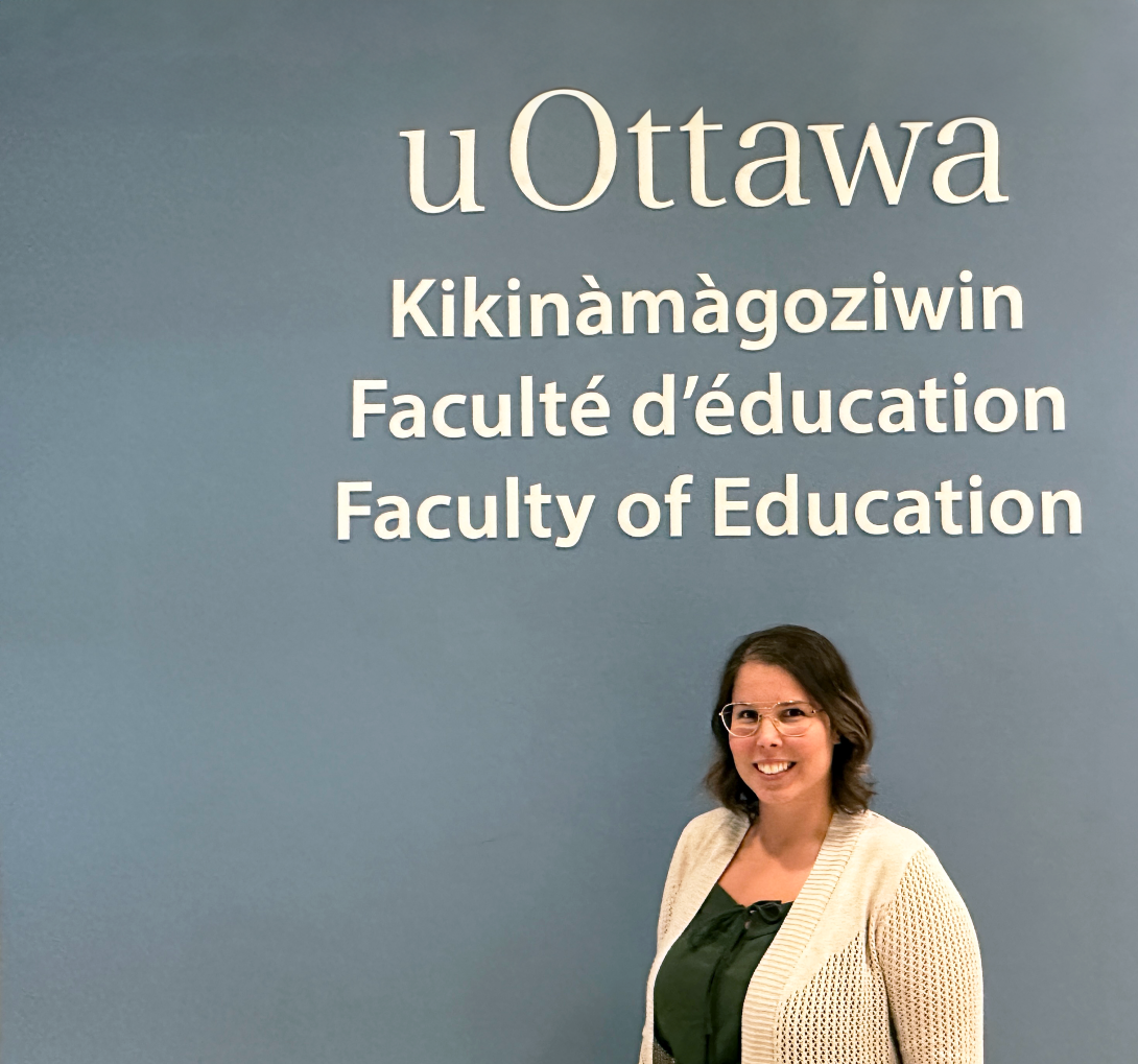 Allison Gagné in front of the Faculty's blue logo wall containing the logo and Faculty of Education in French, English and Annishinabe