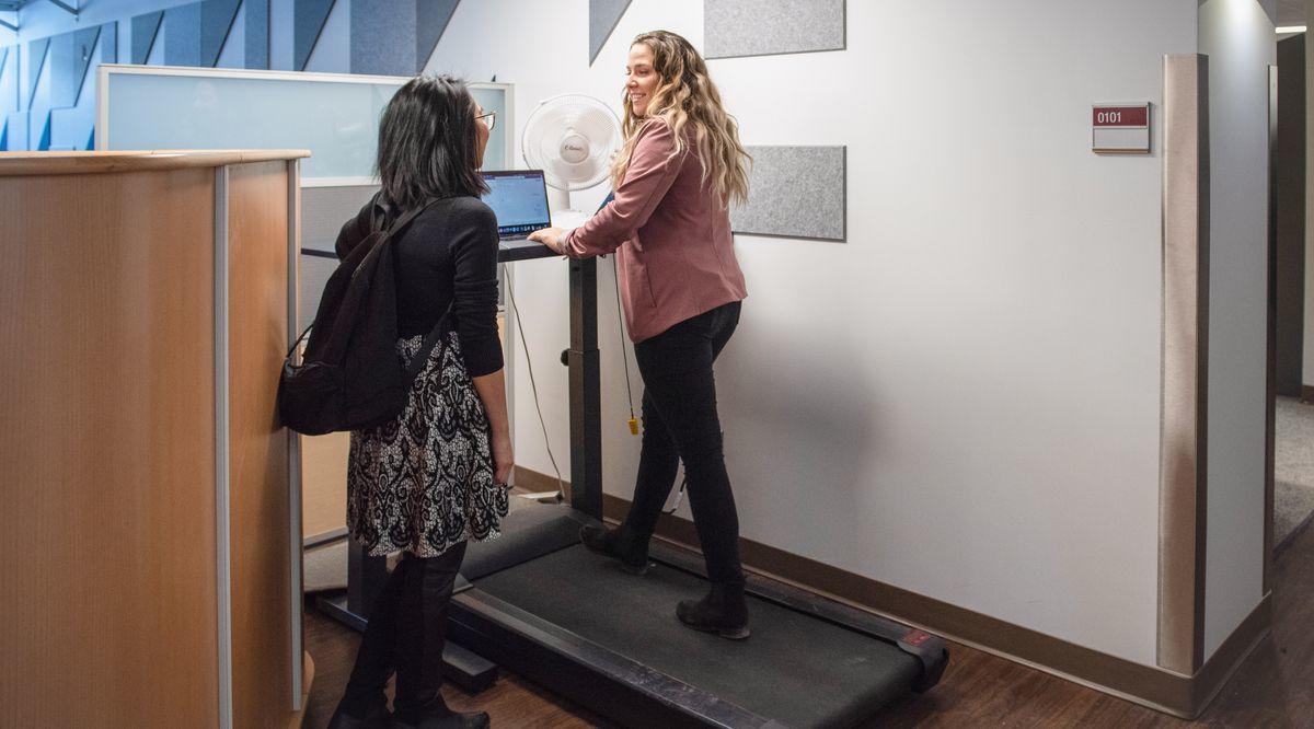 Deux femmes discutent alors que l'une d'entre-elles utilise un bureau de travail avec tapis-roulant.