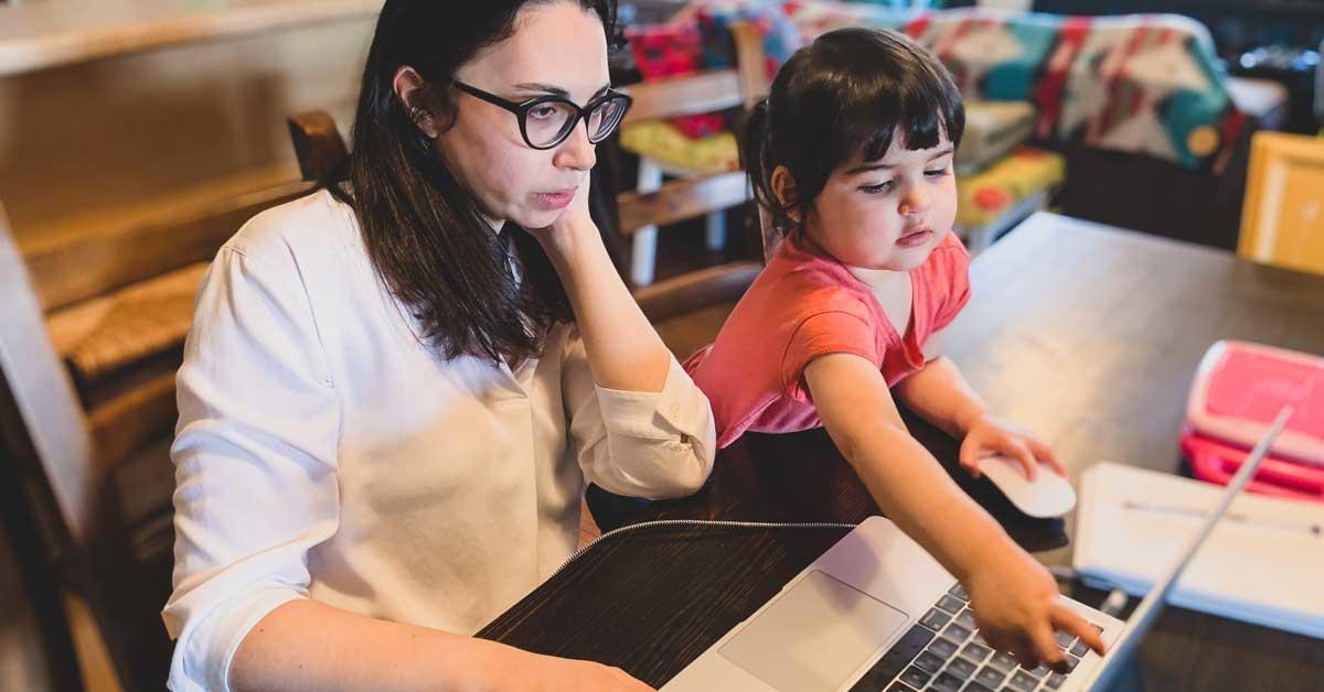 Une femme travaillant sur son ordinateur portable alors que sa fillette est assise à côté d'elle, tient la souris de l'ordinateur et pointe un doigt vers l'écran du portable.