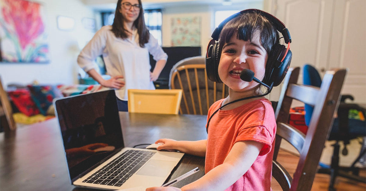 Une une fillette porte des écouteurs sur sa tête. Elle est assise devant un ordinateur portable alors que sa mère l'observe au loin.
