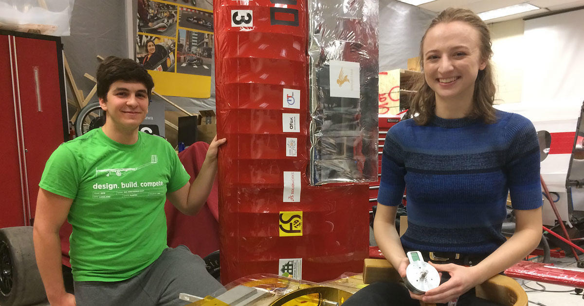 University of Ottawa student Matt Sehri beside a model of an aircraft, holding its wing. University of Ottawa student Sarah Horton sits on a stool, smiling and holding a motor and a curved robotic limb.