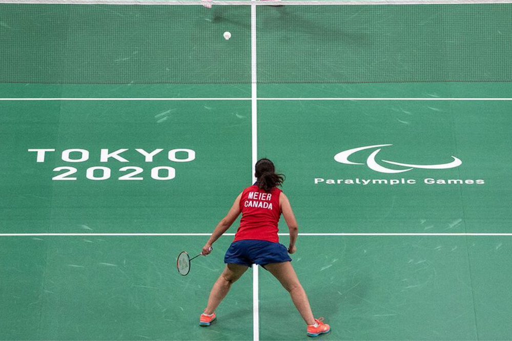 A woman is playing badminton on a green court. The court is marked Tokyo 2020