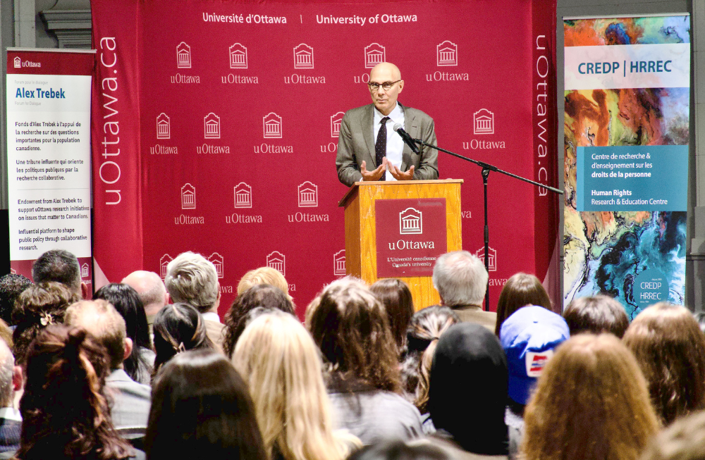  A man stands at a podium on a stage. many people sit facing him.