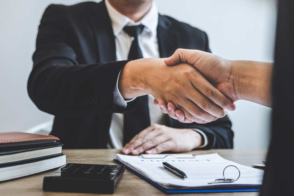 2 people sit across from each other at a desk and shake hands. There is a paper and pen on the desk