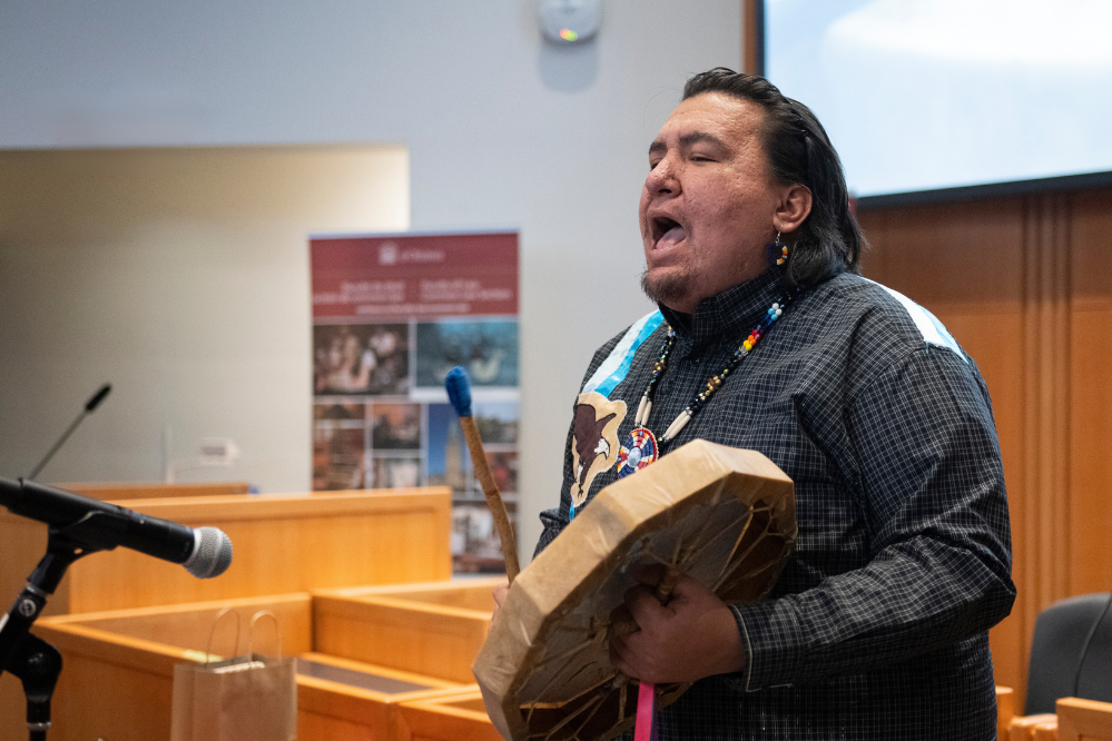 A person holds a drum in one hand and a baton in the other. They appear to be singing.