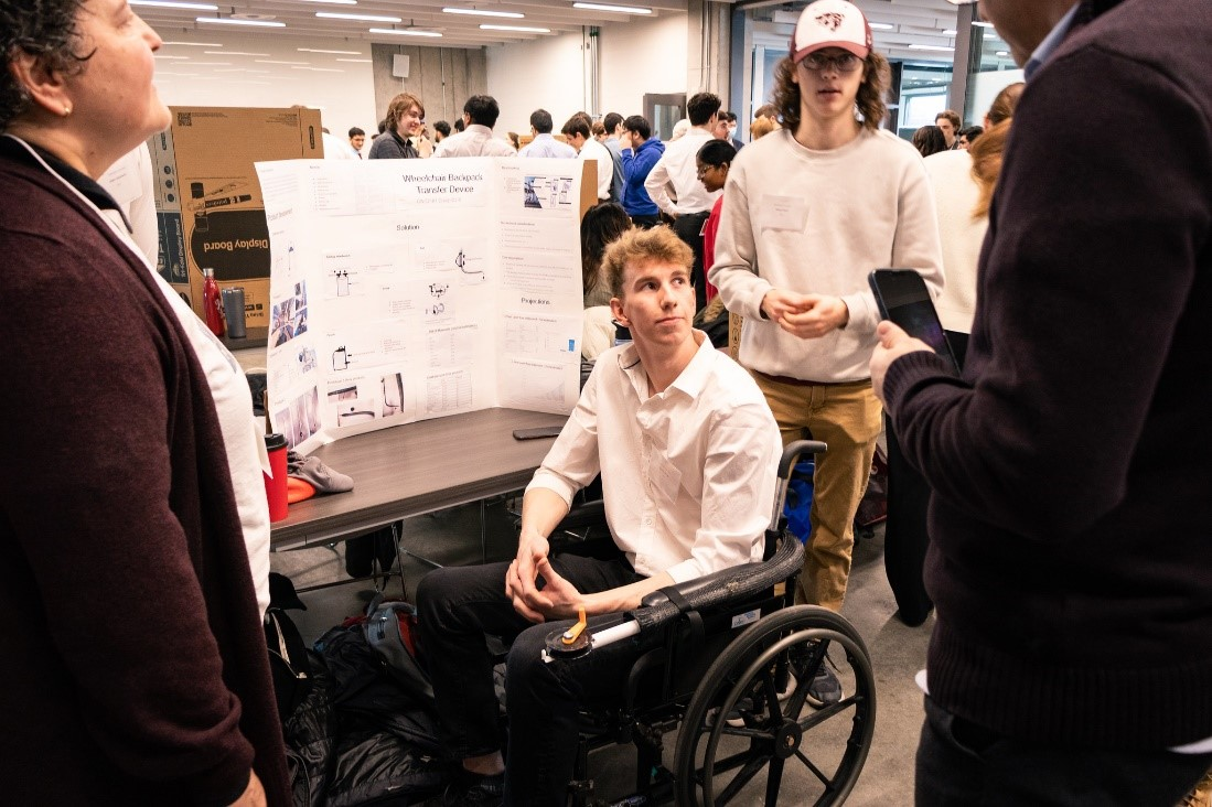Image of a device in prototype stage attached to a wheelchair. The device assists wheelchair users in moving their backpack from behind the chair to a reachable location on the side.