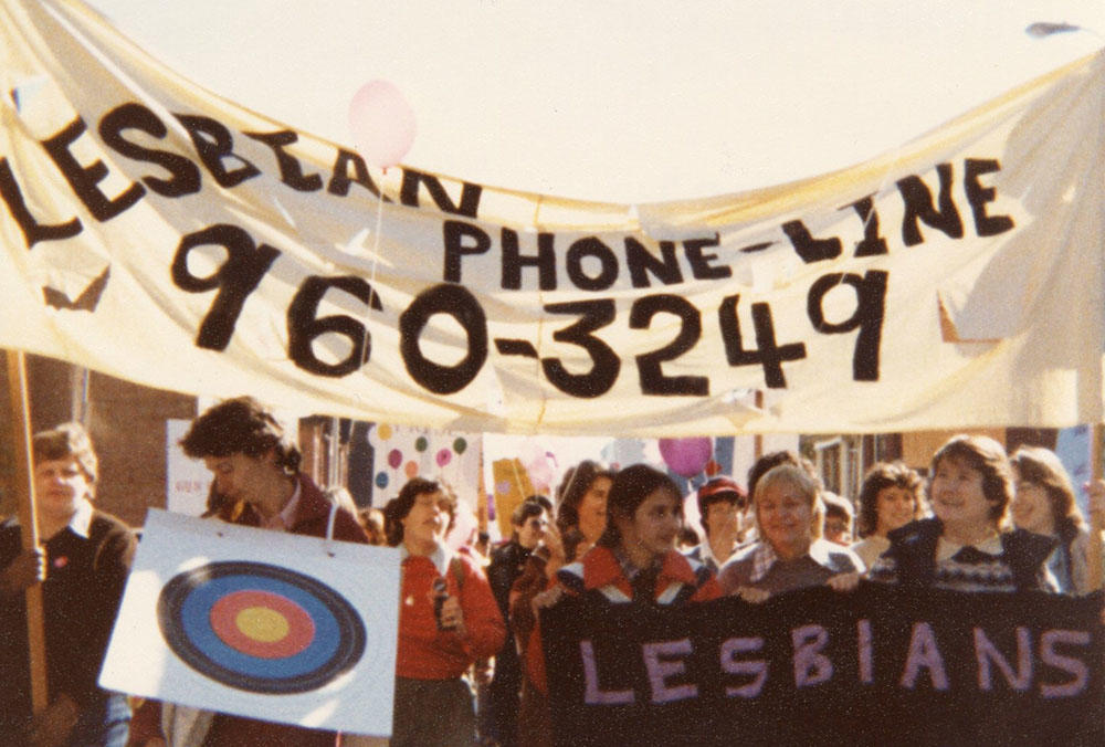 Photographie couleur de femmes tenant des banderoles « Lesbians are everywhere » et « Lesbian phone line 960-3249 » lors d'une manifestation
