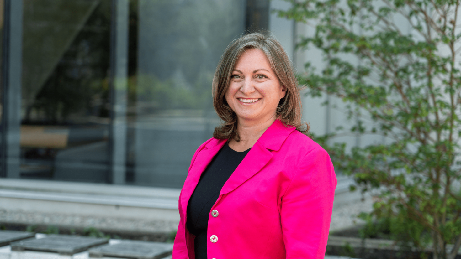 A person in a bright pink blazer stands smiling outdoors in front of a building with large windows and greenery.