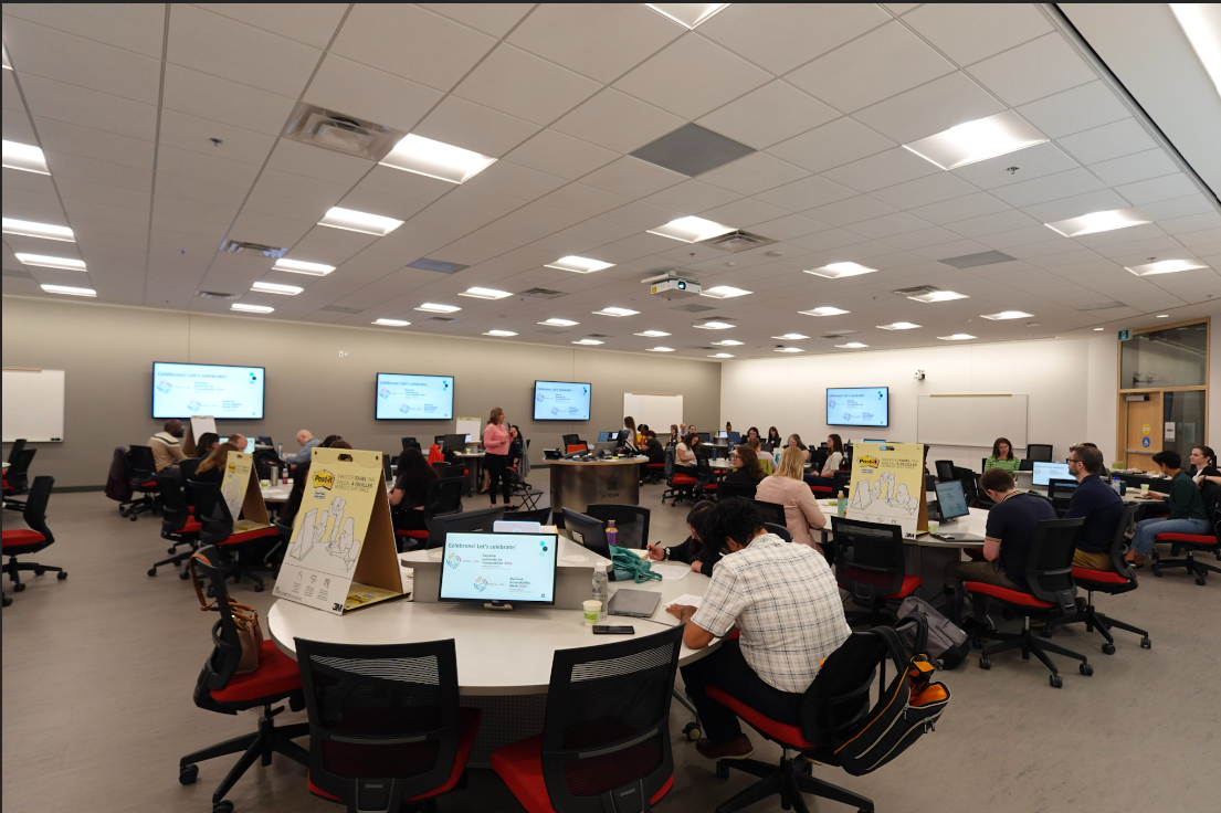 Une salle de classe avec plusieurs personnes assises à des tables, travaillant sur des ordinateurs portables. Trois grands écrans à l'avant affichent des présentations.