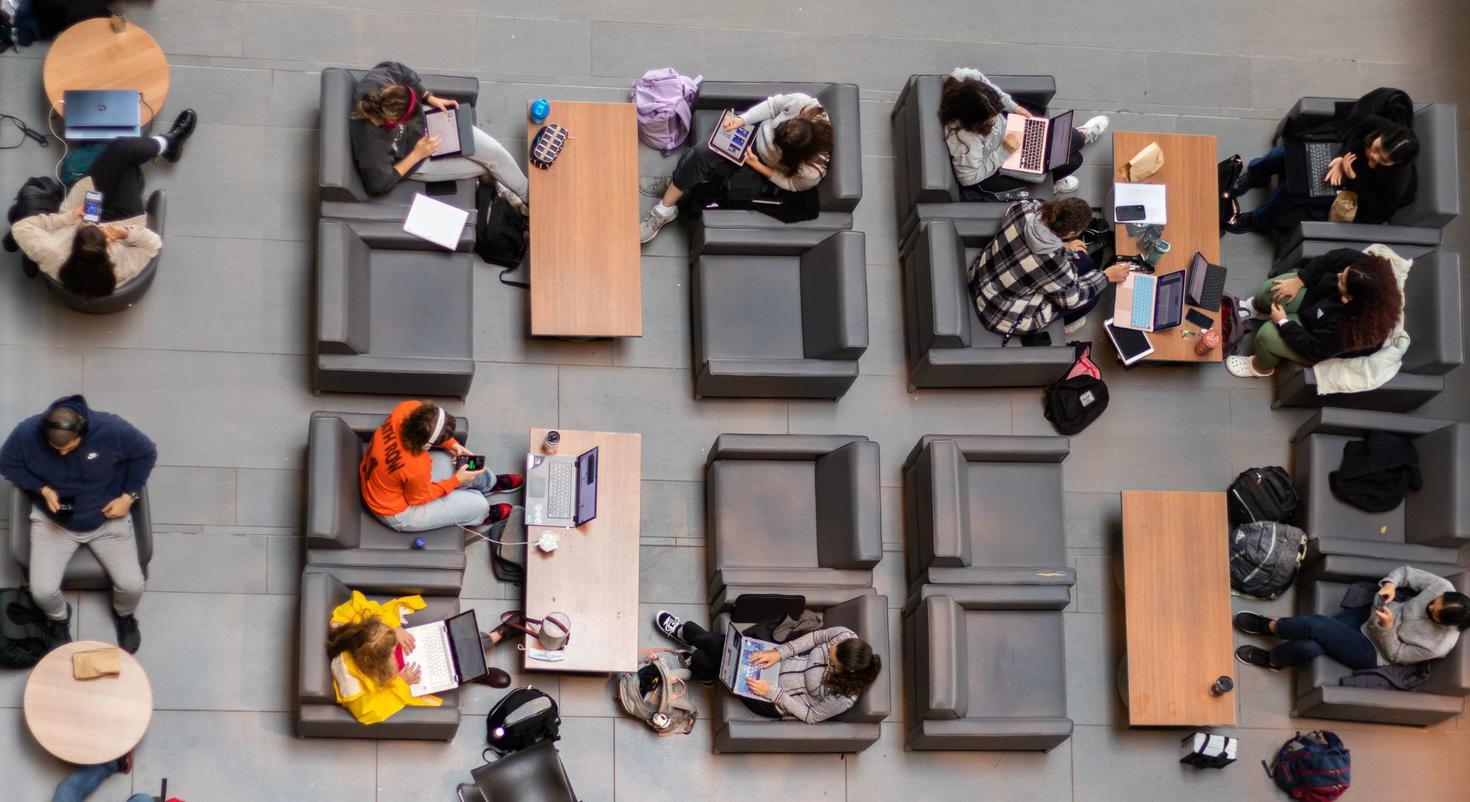 Un groupe d'étudiants assis dans un espace commun, utilisant des ordinateurs portables et des tablettes, assis sur des chaises autour de tables. La scène est vue d'en haut.