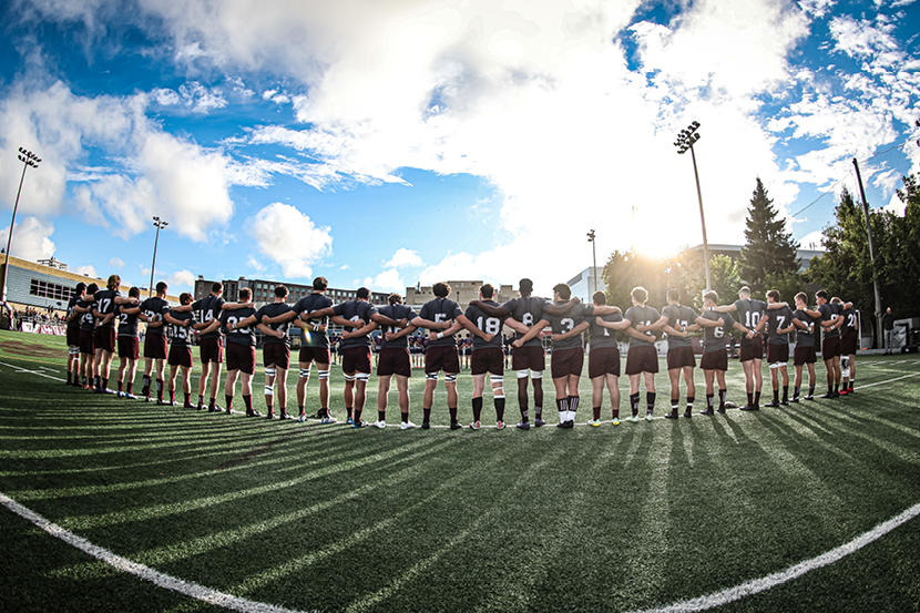 The uOttawa Gee-Gees men's rugby team stand arm in arm in the sunshine on the rugby field.