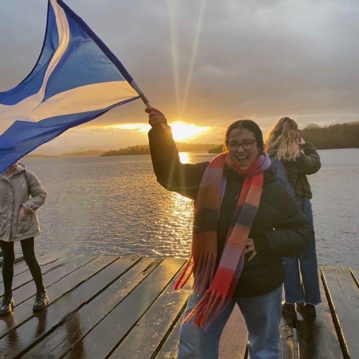 Emma Guzzo waves a Scottish flag as the sun sets across a lake.