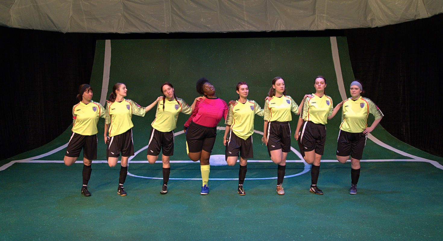 A group of eight soccer players in matching uniforms stands on a green field, striking synchronized poses with one player in a contrasting pink jersey at the center.