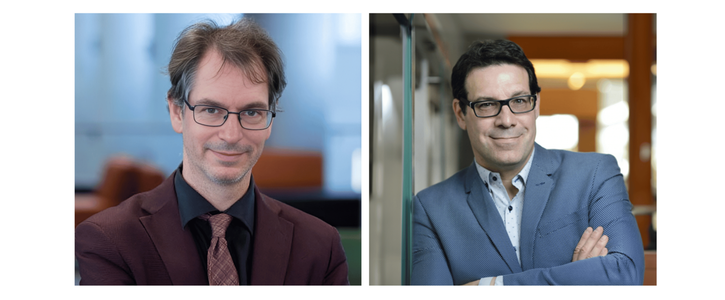 Two men wearing suits and glasses are posing side by side. The man on the left is in a dark suit with a burgundy tie, and the man on the right is in a blue suit with arms crossed.