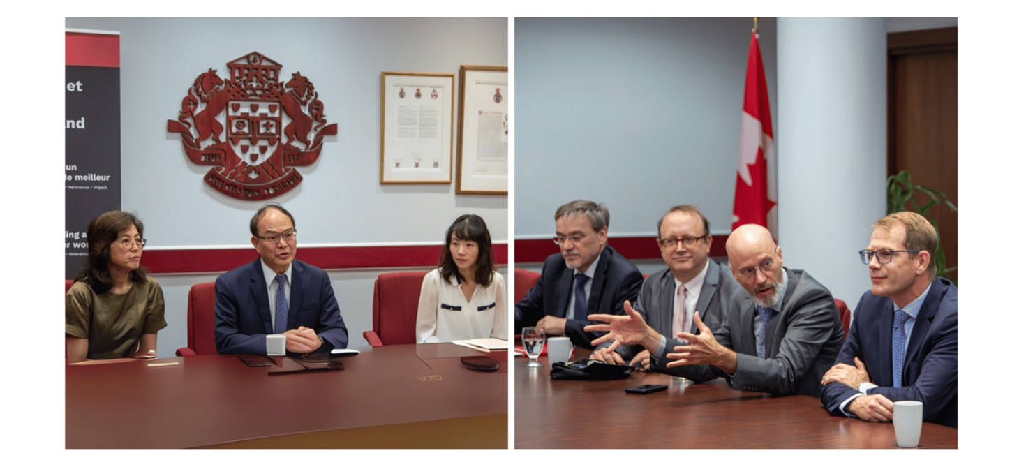 Two groups of people are meeting in a conference room. One side includes three individuals, the other five. A Canadian flag and a coat of arms are visible in the background.