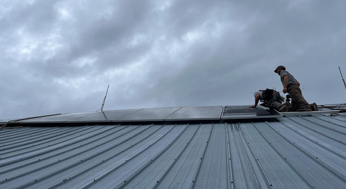 Two workers installing solar panels on a metal roof under a cloudy sky.