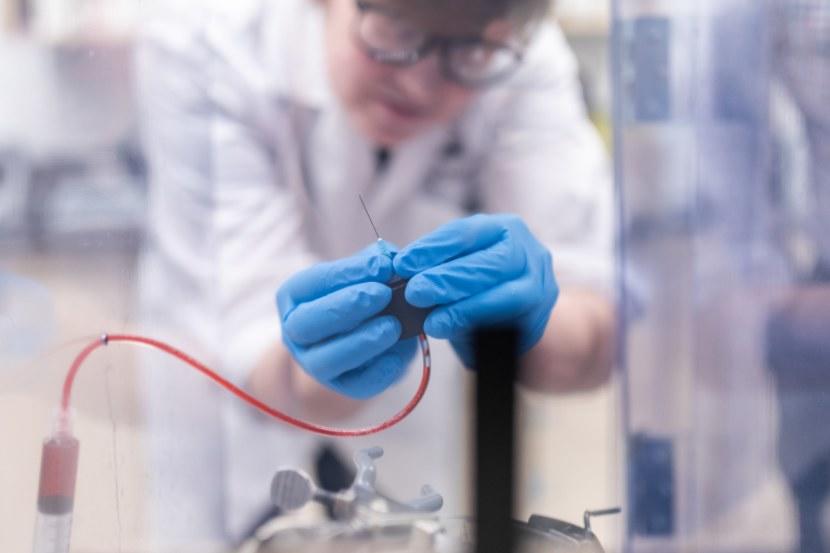 Un chercheur portant une blouse blanche, des lunettes de sécurité et des gants bleus manipule avec précaution une seringue et une tubulure dans un laboratoire.