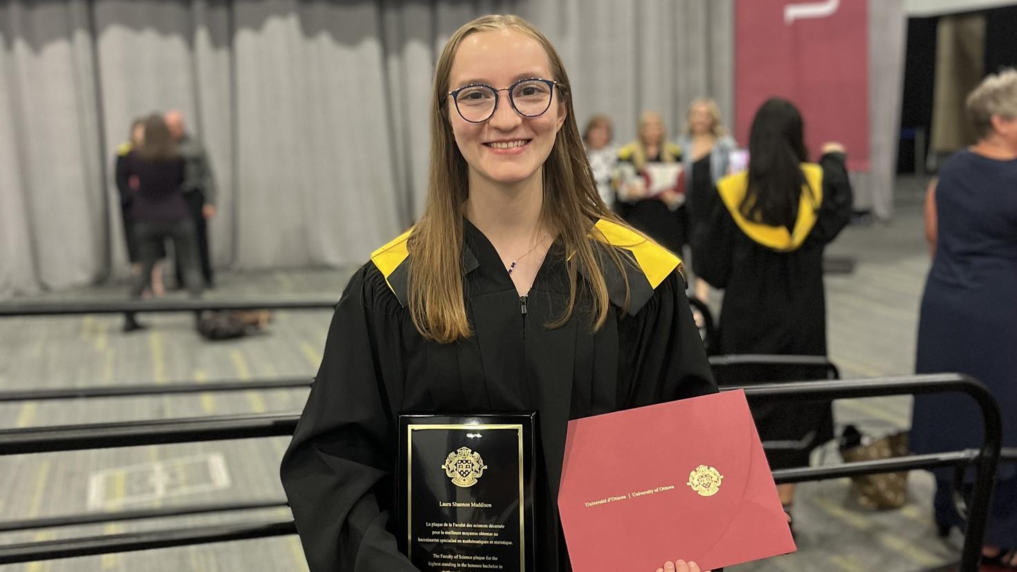 Laura Maddison, vêtue d’une toge noire avec une touche de jaune, se tient debout, devant une barrière, dans la salle des cérémonies de collation des grades