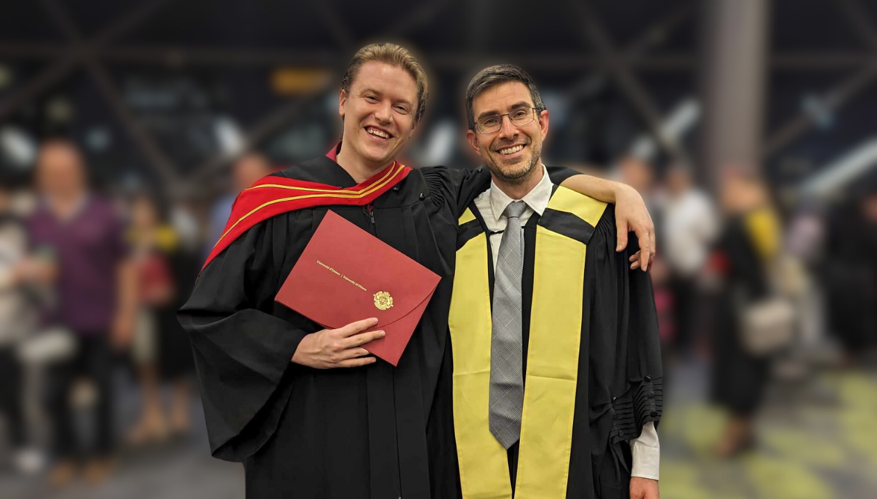 Adam Cook, vêtu d’une toge noire avec une épitoge rouge et or, tenant un diplôme. Le professeur Stephen Newman se tient à sa gauche, vêtu d’une toge noire et d’une écharpe jaune. Ils sont entourés d’autres personnes célébrant l’obtention de leur diplôme. 