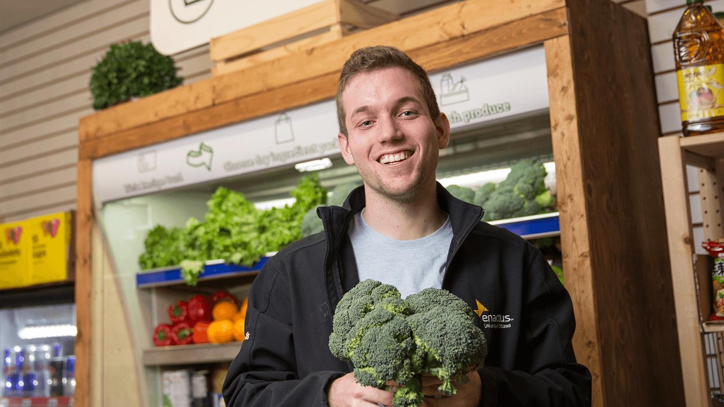 Devant un comptoir réfrigéré plein de produits, Corey Ellis tient en souriant des pieds de brocoli.